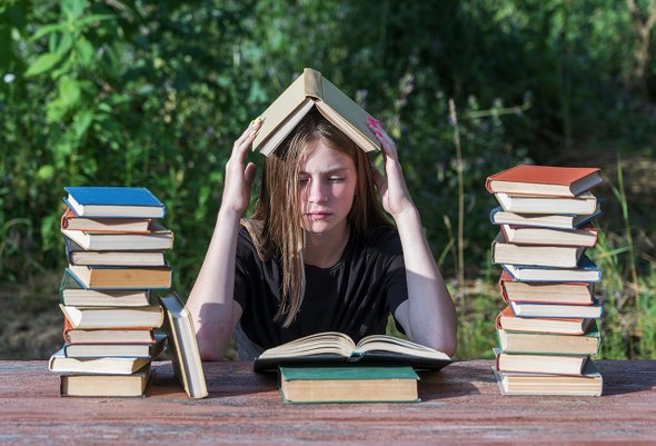 Уставшая девушка подросток читает книгу. Много книг, стопки книг. Подготовка к экзаменам