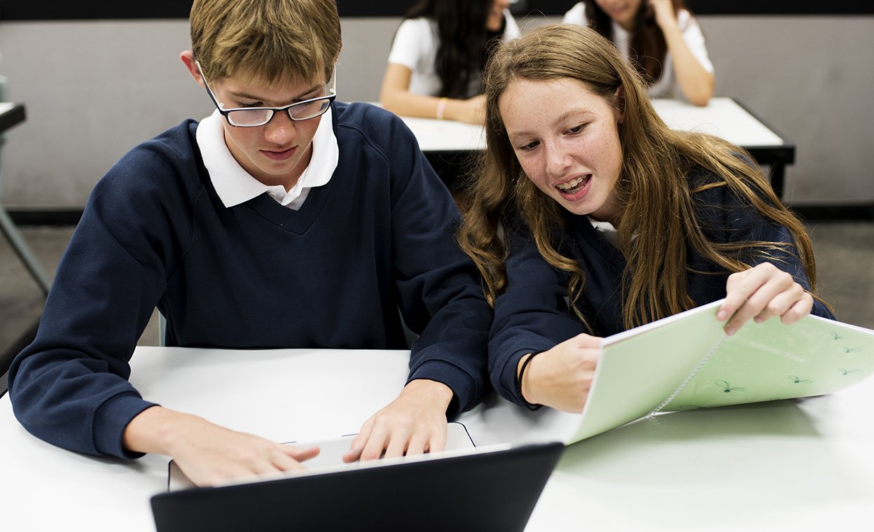 Школьник и школьница сидят за партой с ноутбуком и тетрадями и учатся. Российская и международная программы: как совместить это в школе и не перегрузить ребенка