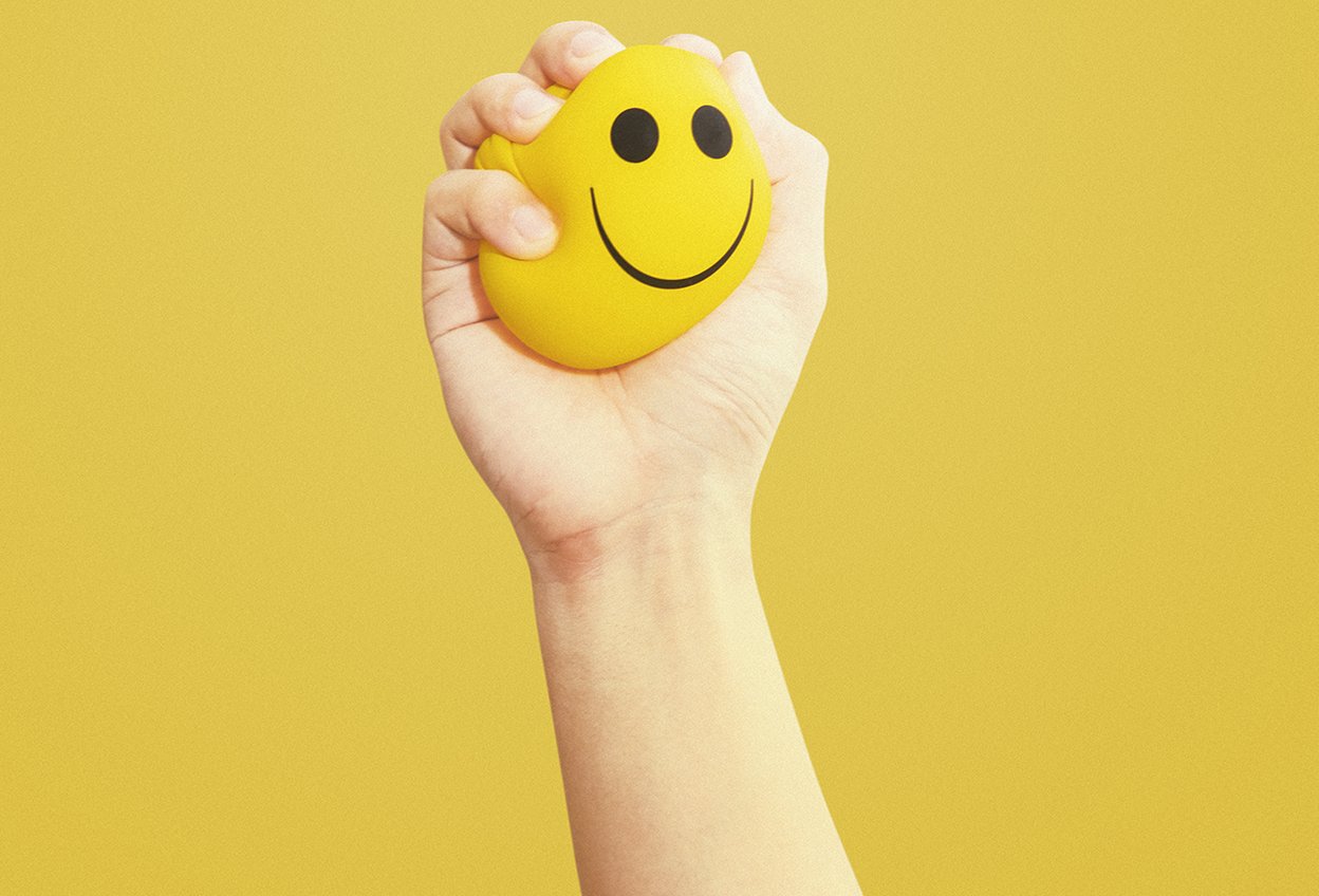 Рука сжимает стресс мячик в виде улыбающегося лица / эмодзи / эмоджи. Стресс и тревога перед экзаменами