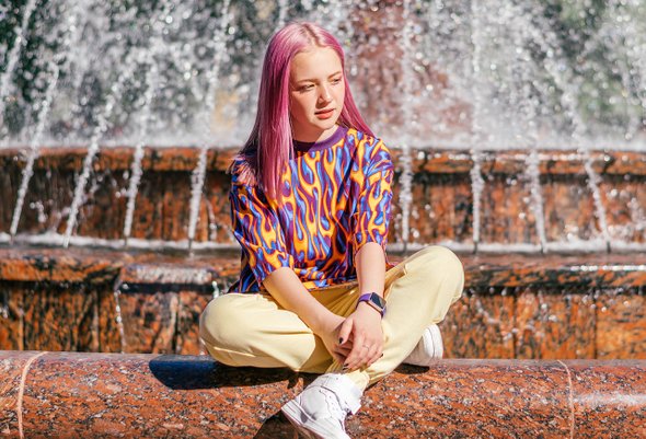 Девочка-подросток с розовыми волосами в яркой одежде на фоне фонтана. Подростковый стиль в одежде